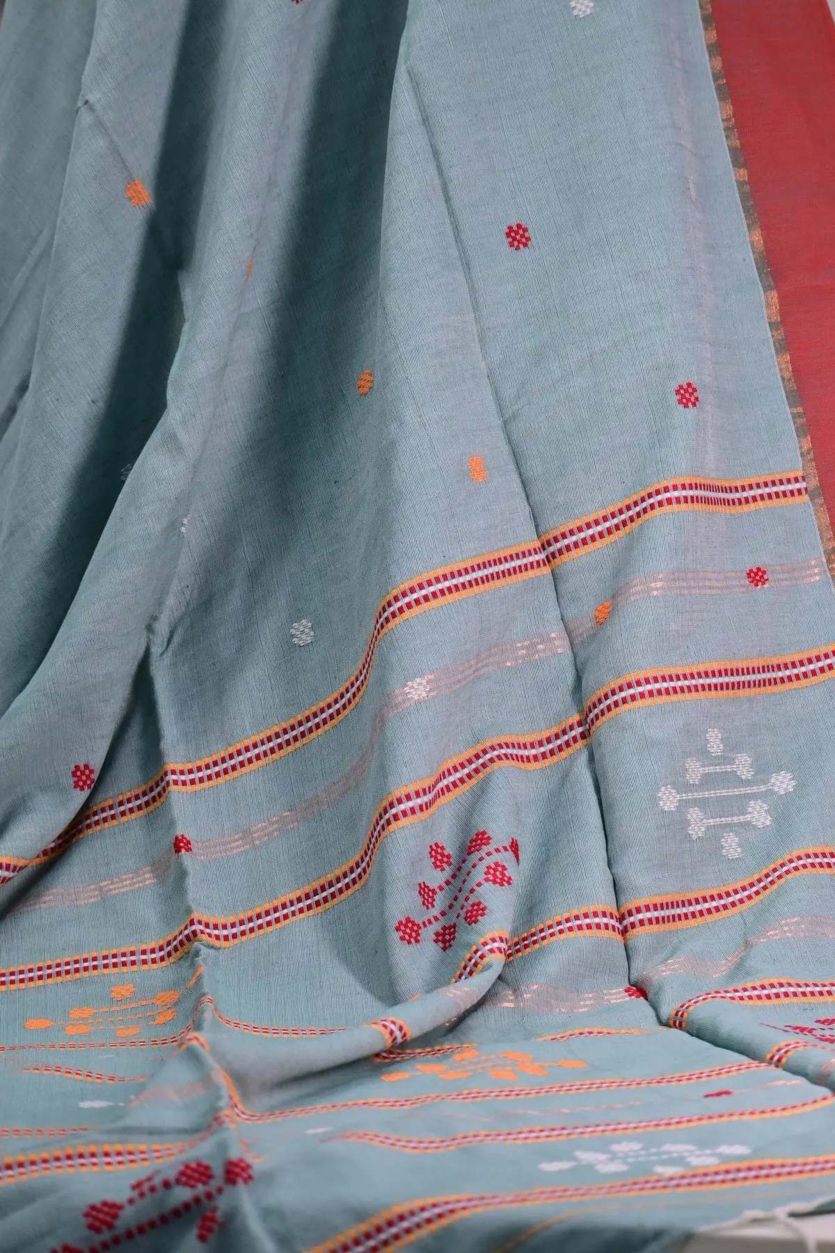 Sage grey-green saree hanging in folds, with red and orange stripes, small square motifs and a coral red border.