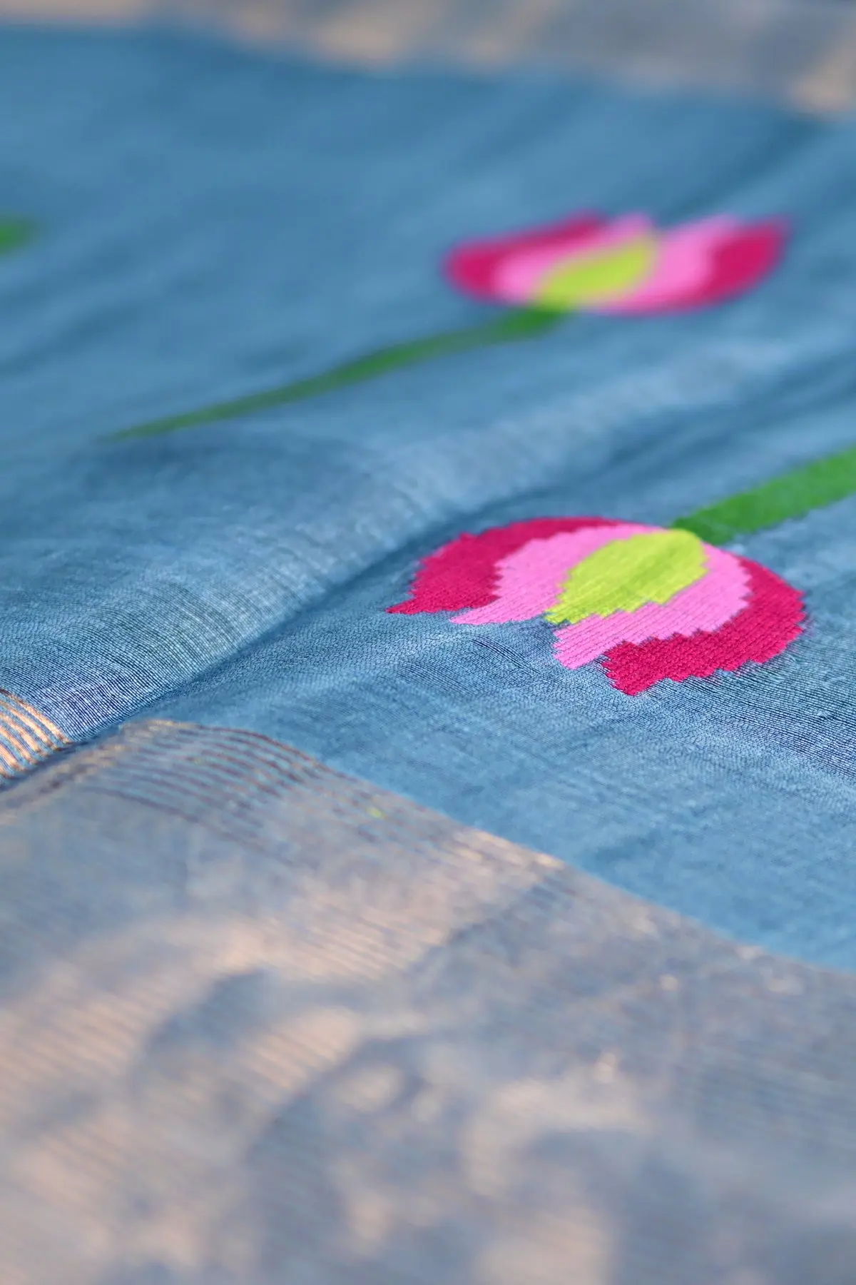 Close-up of a blue linen saree with a pink lotus motif on a green stem beside a copper border.