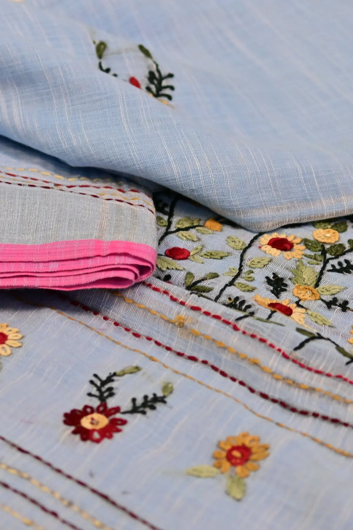 A pale blue linen saree swirled to show dense red, yellow and green floral embroidery.