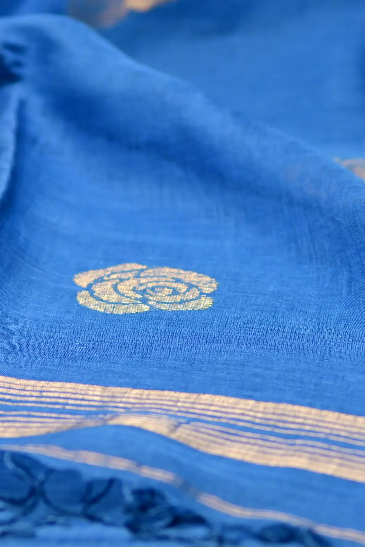 A blue linen saree laid flat, with a gold rose motif below a folded gold and turquoise border.